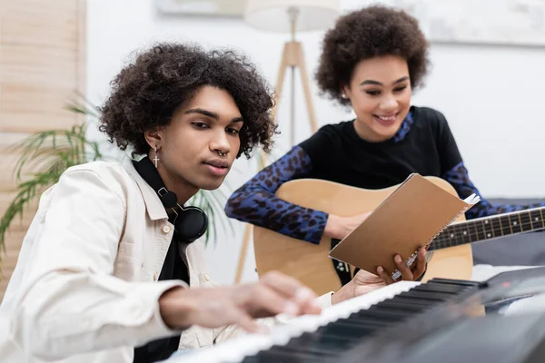 Lächelnde Afroamerikanerin spielt Akustikgitarre neben Freund mit Synthesizer und Notizbuch zu Hause — Stockfoto