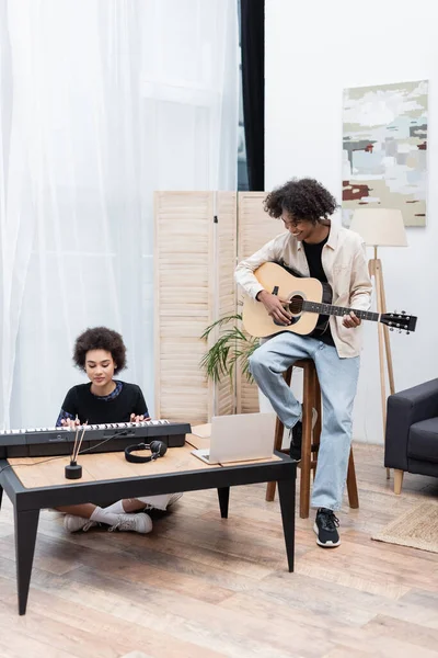 Sorridente uomo afroamericano che suona la chitarra acustica vicino alla fidanzata con sintetizzatore e laptop a casa — Foto stock