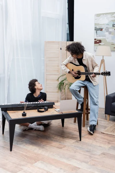 Sorridente coppia afro-americana che si guarda mentre suona strumenti musicali vicino al portatile a casa — Foto stock