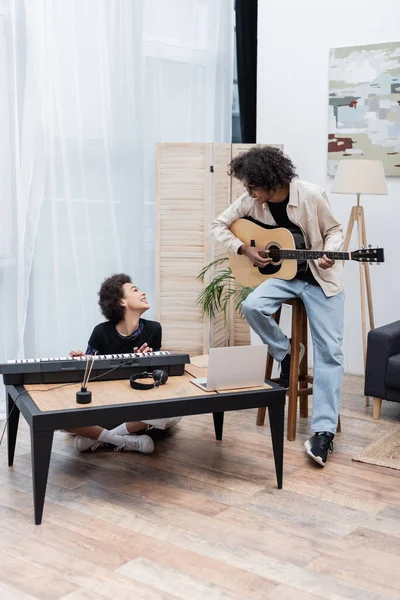 Sonriente pareja afroamericana mirándose mientras toca instrumentos musicales cerca de la computadora portátil en casa - foto de stock