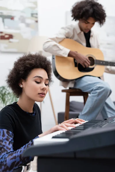 Young african american musician playing synthesizer near blurred boyfriend with acoustic guitar at home — Stock Photo