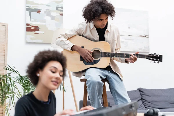 African american man playing acoustic guitar near blurred girlfriend with synthesizer at home — Stock Photo