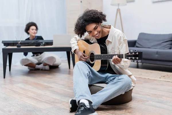 Alegre homem americano africano tocando guitarra acústica perto de namorada turva em casa — Fotografia de Stock