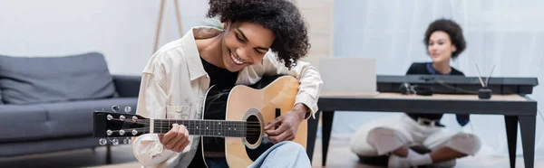 Young african american man playing acoustic guitar near blurred girlfriend in living room, banner — Stock Photo