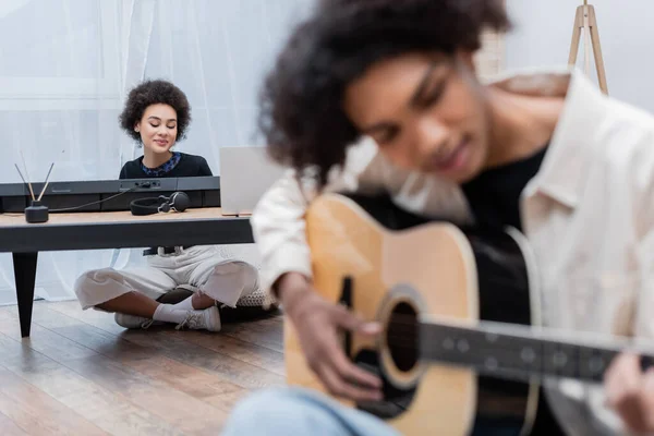 Sorridente donna afroamericana seduta vicino al computer portatile, sintetizzatore e fidanzato sfocato che suona la chitarra acustica a casa — Foto stock