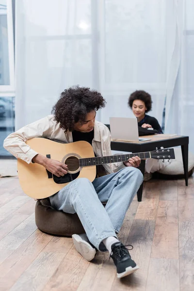 Hombre afroamericano tocando la guitarra acústica cerca de novia borrosa y portátil en casa - foto de stock