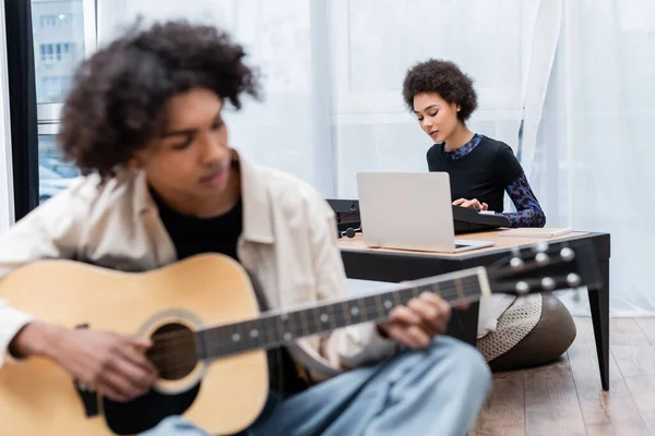 Africano americano mulher jogando sintetizador perto laptop e namorado borrado com guitarra acústica em casa — Fotografia de Stock