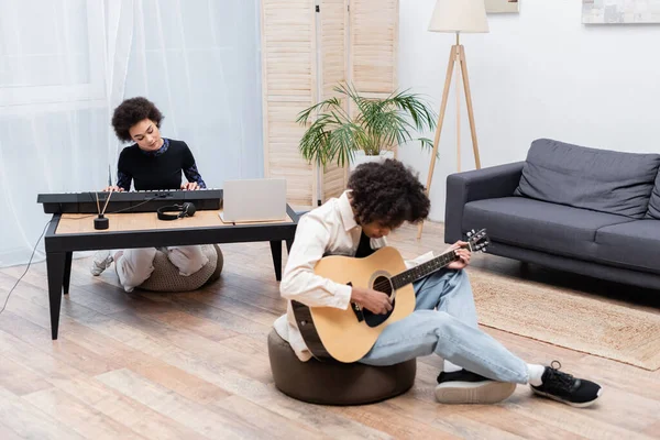 African american woman playing synthesizer near laptop and boyfriend with acoustic guitar at home — Stock Photo