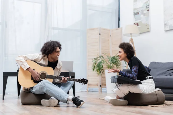 Alegre mujer afroamericana con sintetizador hablando con novio con guitarra acústica en casa - foto de stock