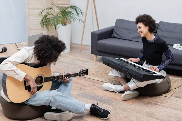 Musicien afro-américain souriant jouant du synthétiseur près du petit ami avec guitare acoustique à la maison — Photo de stock