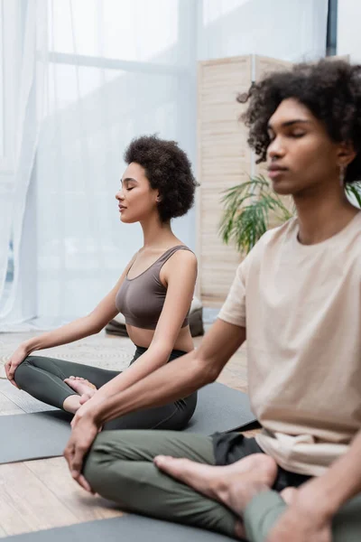 Africano americano mulher sentada em ioga pose perto de namorado borrado em casa — Fotografia de Stock