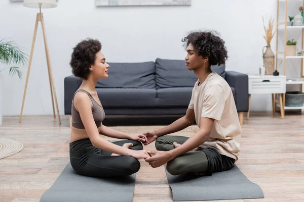 Seitenansicht eines jungen afrikanisch-amerikanischen Paares, das Händchen hält, während es zu Hause auf Matten meditiert — Stockfoto