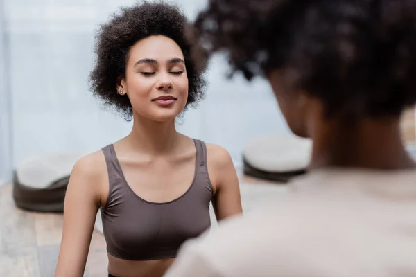 Femme afro-américaine avec les yeux fermés pratiquant le yoga près de copain flou à la maison — Photo de stock