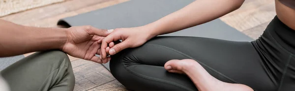 Vista recortada de una pareja afroamericana cogida de la mano mientras practica yoga en casa, pancarta - foto de stock