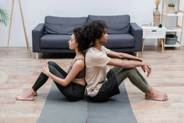 Vue latérale du jeune couple afro-américain méditant dos à dos sur des tapis de yoga à la maison — Photo de stock
