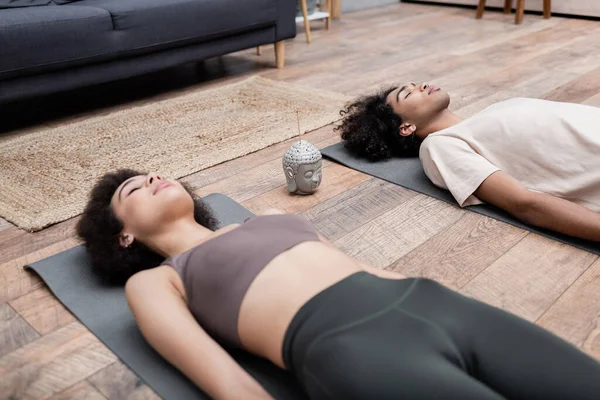 Joven mujer afroamericana acostada en una esterilla de yoga cerca de un palo de incienso y su novio meditando en casa - foto de stock