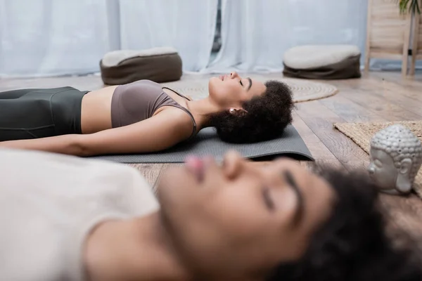 Giovane donna afroamericana in abbigliamento sportivo meditando vicino fidanzato offuscata a casa — Foto stock