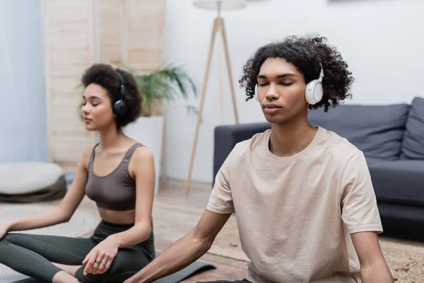 African american man in headphones meditating near girlfriend at home — Stock Photo