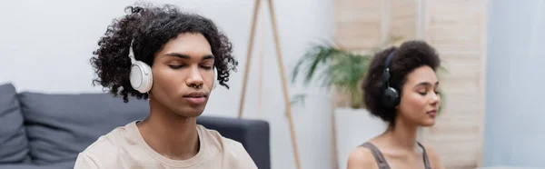 Junge afrikanisch-amerikanische Mann mit Kopfhörern meditiert in der Nähe verschwommene Freundin zu Hause, Banner — Stockfoto