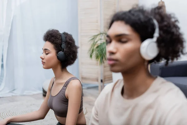 African american woman in headphones practicing yoga near blurred boyfriend at home — Stock Photo
