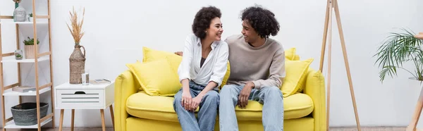 Positivo casal afro-americano olhando um para o outro no sofá em casa, bandeira — Fotografia de Stock