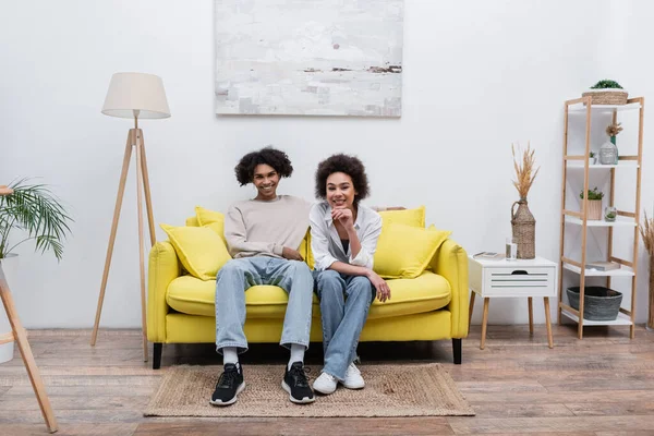 Sonriente mujer afroamericana mirando a la cámara cerca de novio en el sofá en casa - foto de stock