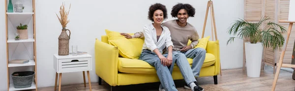 Joyeux couple afro-américain regardant la caméra sur le canapé près du chevalet à la maison, bannière — Photo de stock