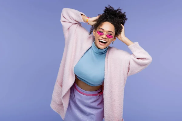 Щаслива афроамериканська жінка в стильних сонцезахисних окулярах, що регулюють кучеряве волосся ізольовано на фіолетовому — стокове фото