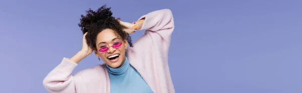 Heureuse femme afro-américaine dans des lunettes de soleil élégantes ajustant les cheveux bouclés isolés sur violet, bannière — Photo de stock
