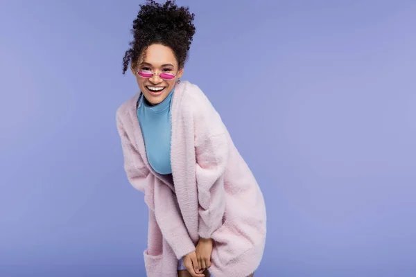 Счастливая африканская американка в стильных солнцезащитных очках позирует изолированно на фиолетовой — стоковое фото