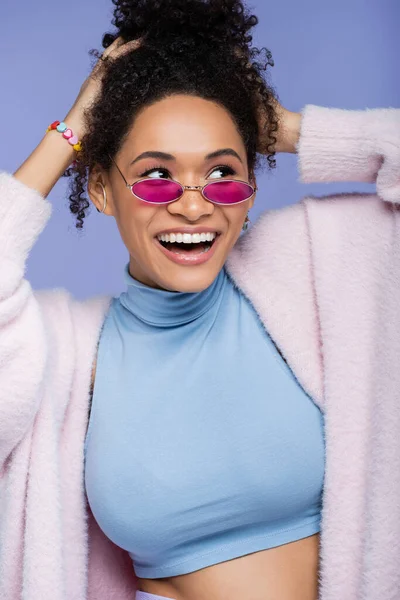 Femme afro-américaine gaie en lunettes de soleil roses ajustant les cheveux bouclés isolés sur violet — Photo de stock