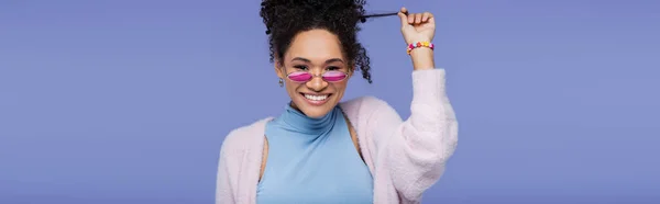 Joyeuse femme afro-américaine en lunettes de soleil roses tirant les cheveux bouclés isolés sur violet, bannière — Photo de stock