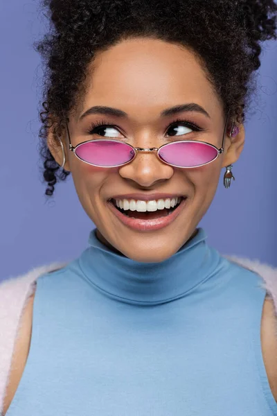 Крупним планом позитивна афроамериканка в рожевих сонцезахисних окулярах ізольована на фіолетовому — стокове фото