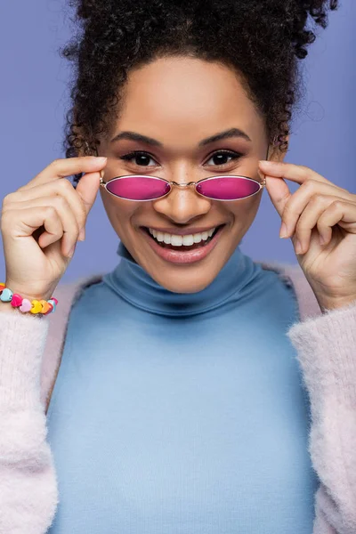 Mulher americana africana feliz ajustando óculos de sol elegantes isolados em violeta — Fotografia de Stock