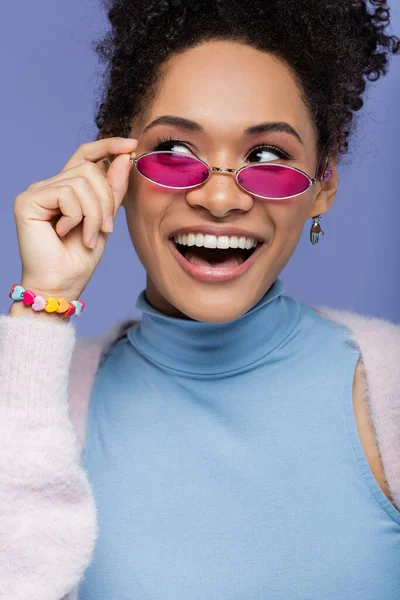 Щаслива афроамериканка, що налаштовує модні сонцезахисні окуляри ізольовані на фіолетовому — стокове фото