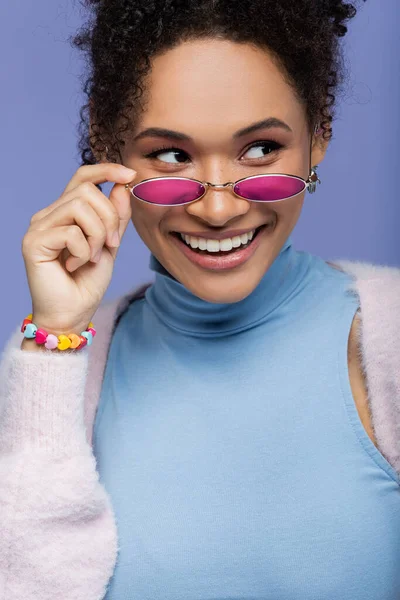 Позитивна афроамериканська жінка регулює модні сонцезахисні окуляри ізольовані на фіолетовому — стокове фото