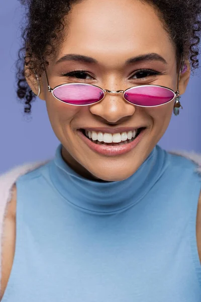 Зблизька приємна американка в рожевих сонцезахисних окулярах посміхається ізольовано на фіолетовому — стокове фото