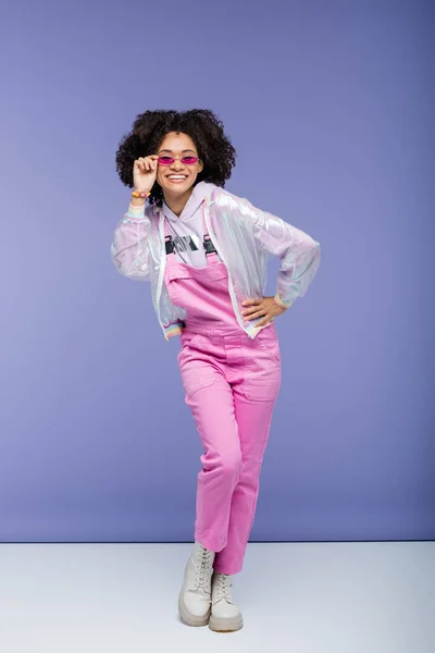 Mujer afroamericana feliz en mono rosa ajustando gafas de sol elegantes mientras posando con la mano en la cadera en púrpura — Stock Photo