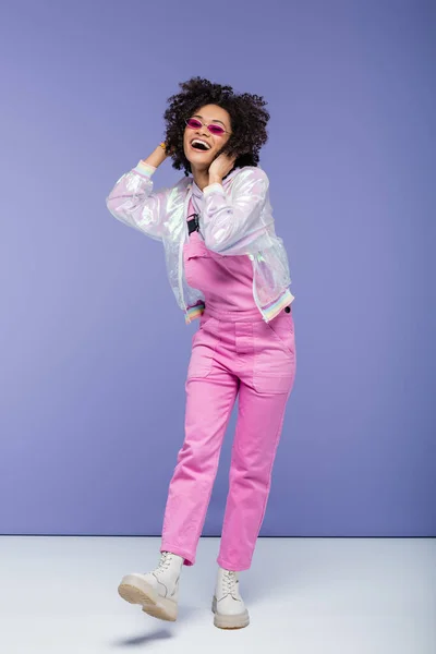 Pleine longueur de femme afro-américaine excitée dans des lunettes de soleil élégantes et salopettes roses ajuster les cheveux bouclés sur violet — Photo de stock