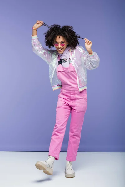 Full length of positive african american woman in sunglasses and pink overalls pulling curly hair on purple — Stock Photo