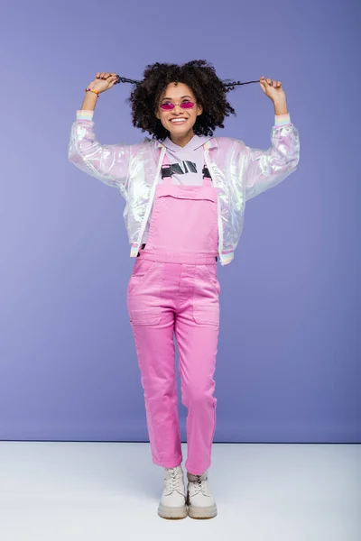 Full length of happy african american woman in pink sunglasses and overalls pulling curly hair on purple — Stock Photo