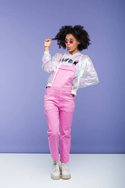 Full length of african american woman in pink sunglasses and overalls pulling curly hair and posing with hand on hip on purple — Stock Photo