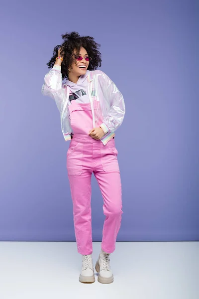 Full length of smiling african american woman in sunglasses and overalls adjusting curly hair on purple — Stock Photo