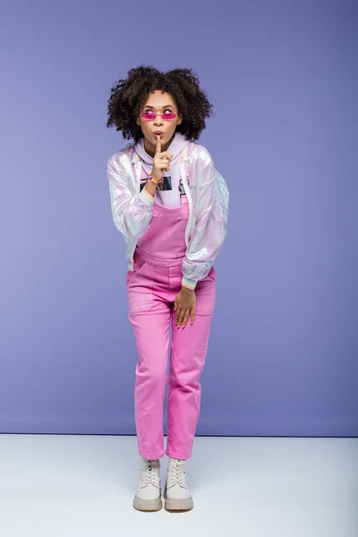 Longitud completa de la mujer afroamericana en gafas de sol y overoles mostrando signo de silencio en púrpura - foto de stock