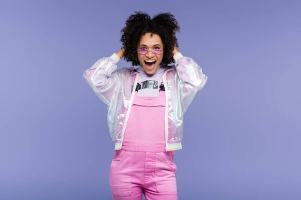 Excited african american woman in stylish sunglasses and overalls adjusting curly hair isolated on purple — Stock Photo