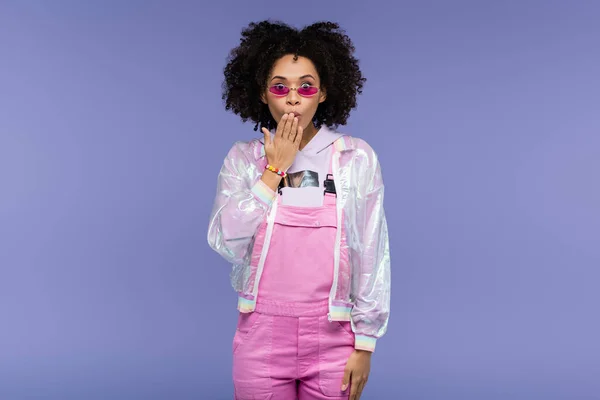 Surprised african american woman in stylish sunglasses covering mouth isolated on purple — Stock Photo