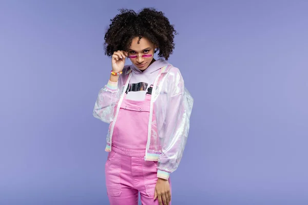 Curly african american woman in pink overalls adjusting sunglasses isolated on purple — Stock Photo