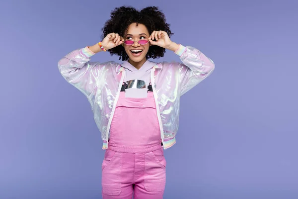 Счастливая африканская американская молодая женщина в розовом комбинезоне регулирует солнцезащитные очки, изолированные на фиолетовый — стоковое фото