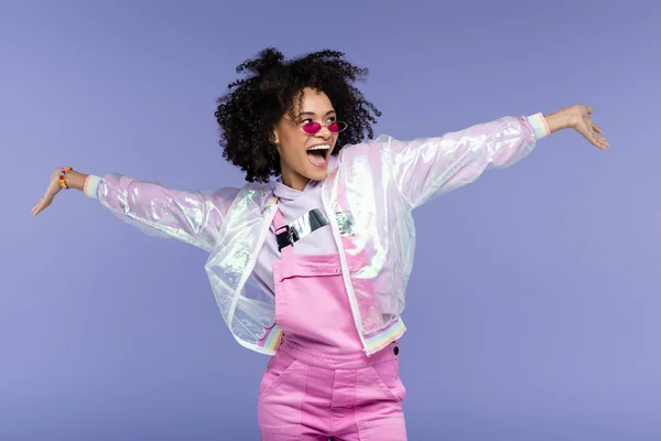 Femme afro-américaine étonnante dans des lunettes de soleil élégantes debout avec les mains tendues isolées sur violet — Photo de stock