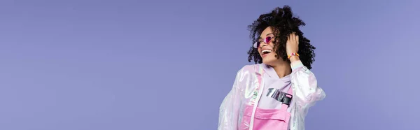 Счастливая африканская американка в стильных солнцезащитных очках регулирует волосы, позируя изолированно на фиолетовом, баннере — стоковое фото